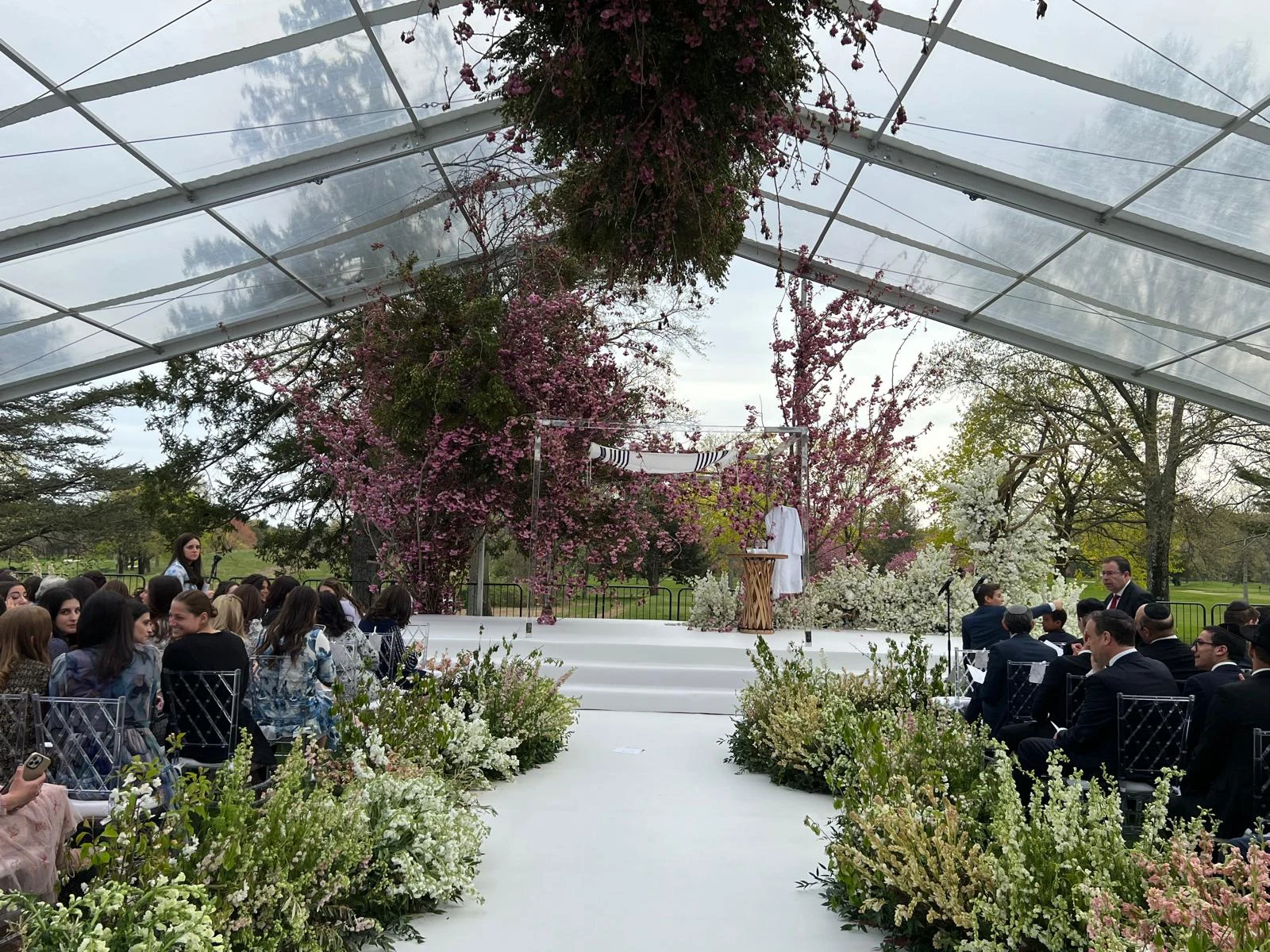 Floral chuppah ceremony under a clear-top tent surrounded by cherry blossoms