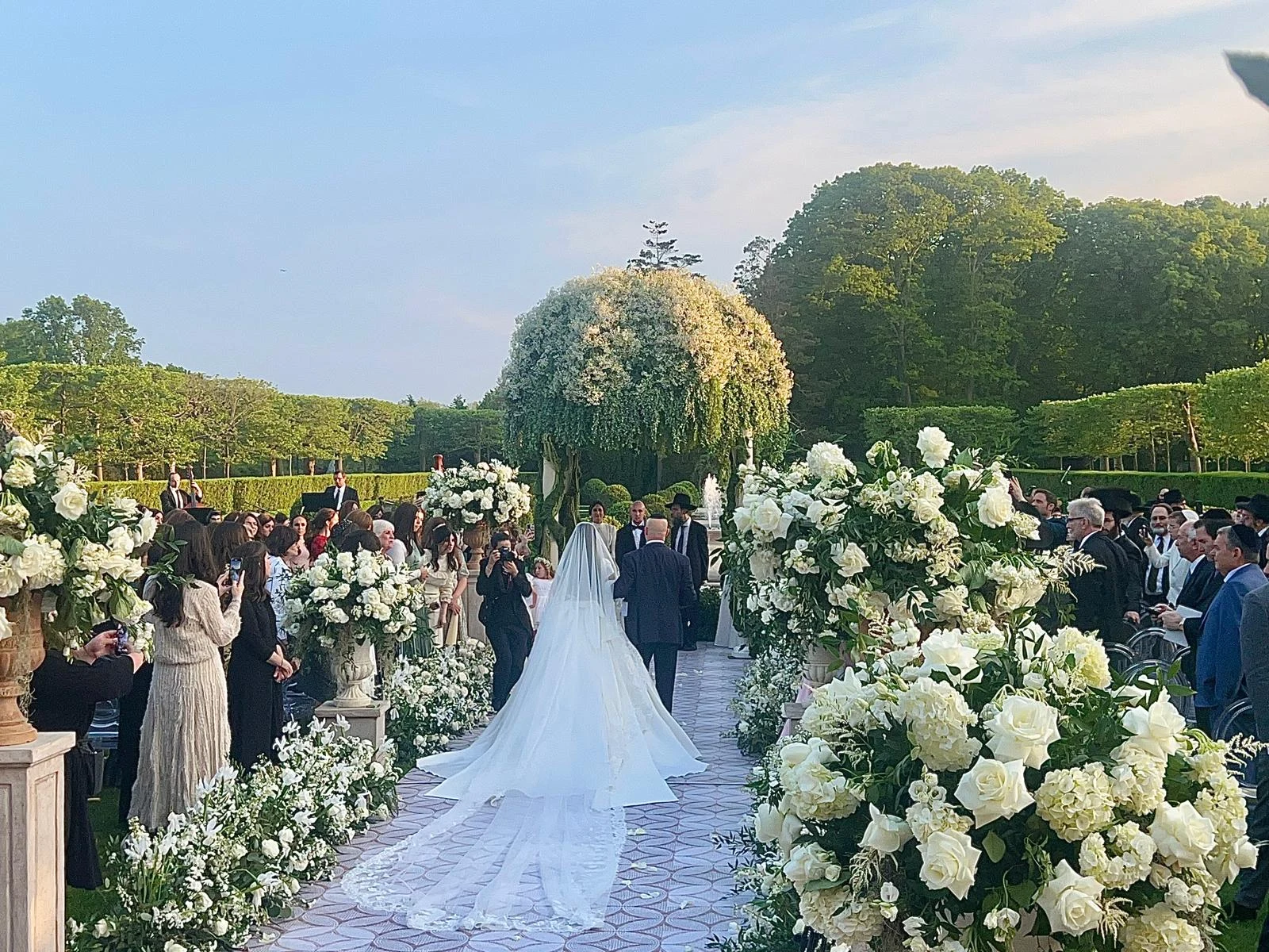 Bride walking down a floral-lined aisle toward a lush garden chuppah